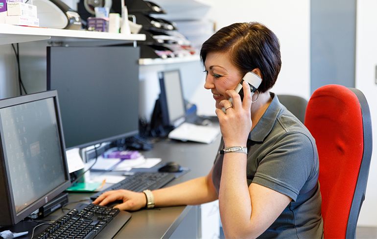 Eine Apothekerin der Petrisberg Apotheke sitzt im Büro und arbeitet mit dem PC, gleichzeitig führt sie ein Telefonat.