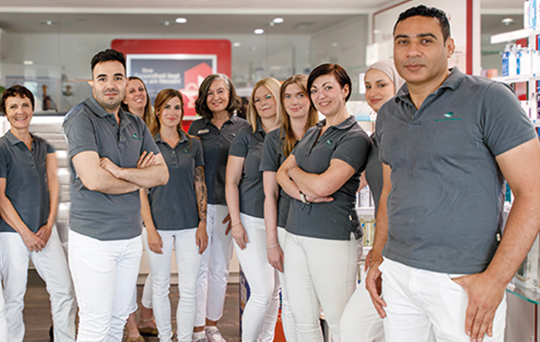 Das Team der Petrisberg Apotheke in Trier steht in der Apotheke von den Medikamentenregalen zusammen.